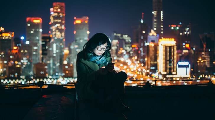 A woman looks at an LED mobile phone screen in front of a cityscape at night